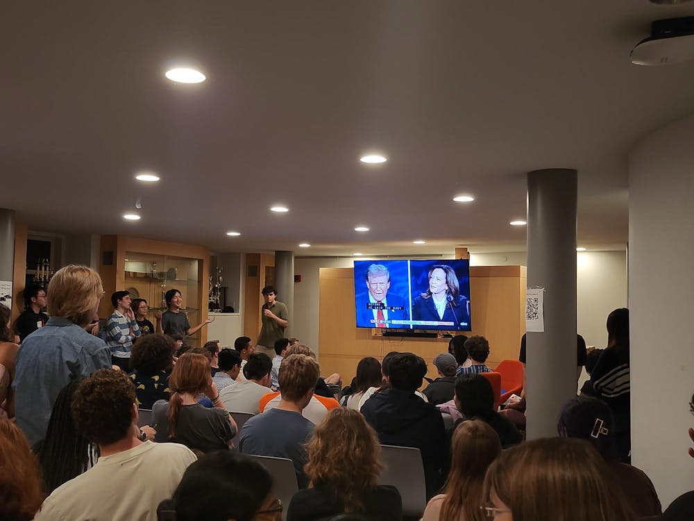 A crowd of mostly seated people faces away from the camera and towards a TV with a split screen showing two people in suits