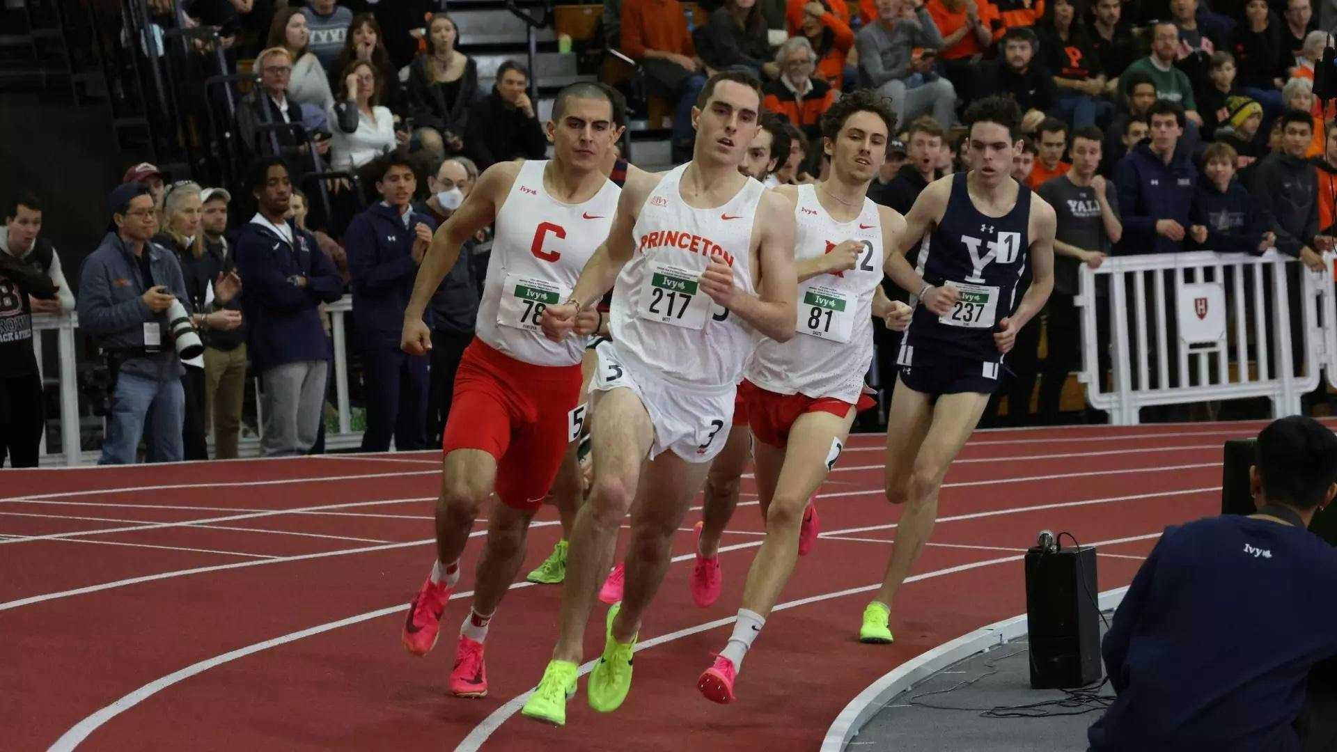 Man in white jersey and neon green sneakers runs on a track ahead of some other runners.