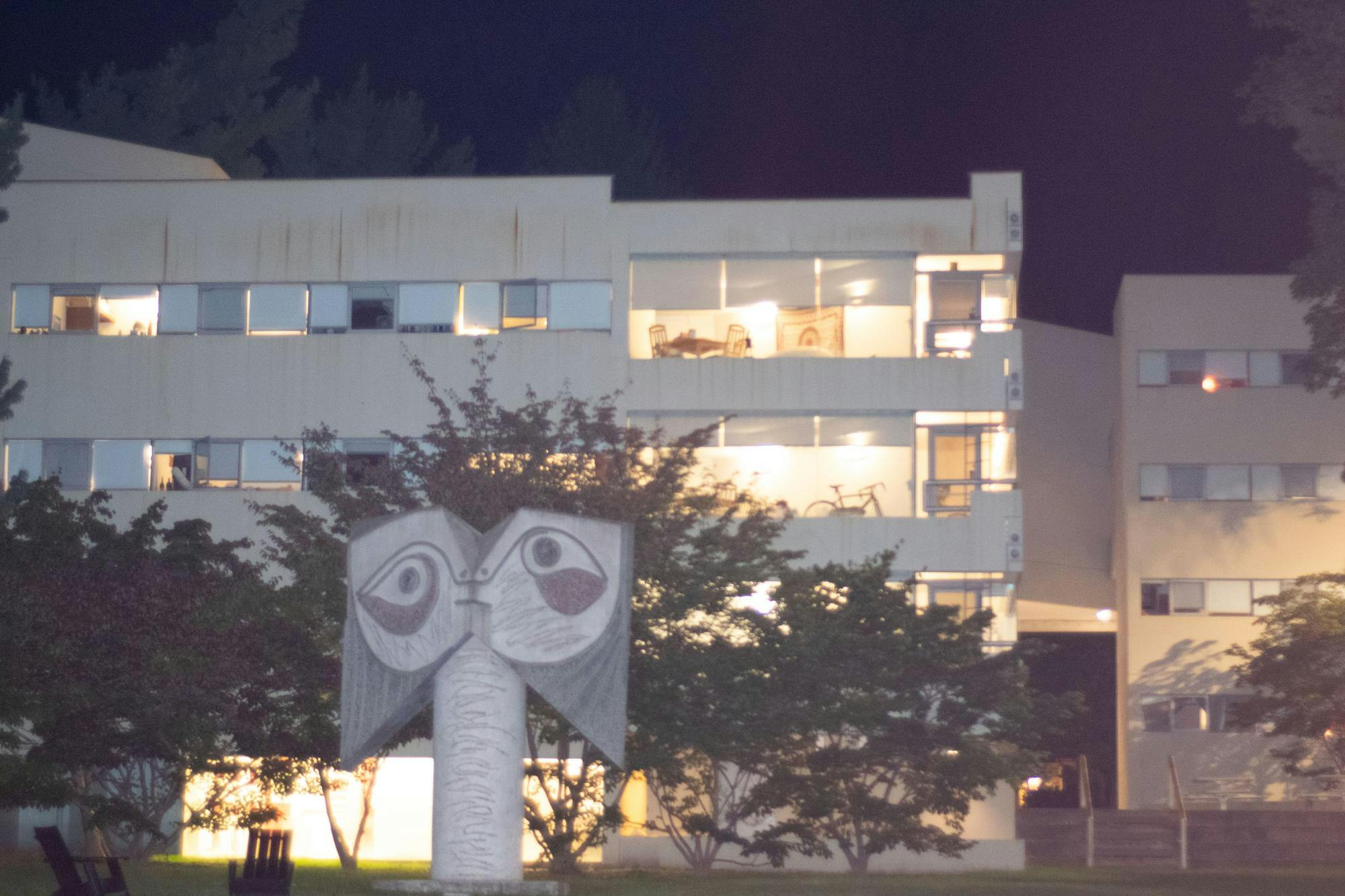 A modern apartment building stands behind a statue in the style of a totem pole. The sky is dark.