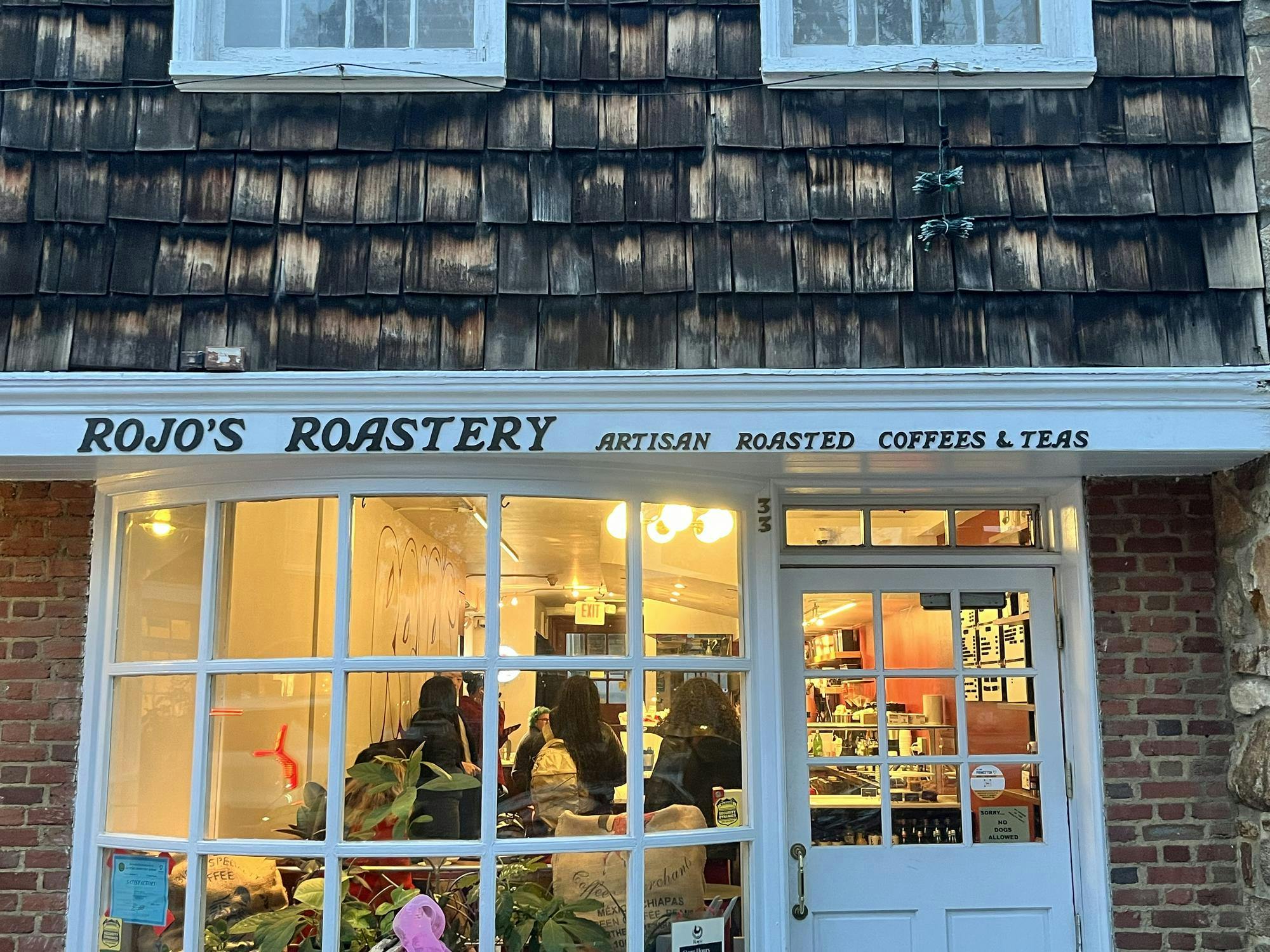 Front view of a coffee shop with people inside. On the white trim, it says "Rojo's Roastery Artisan Roasted Coffees & Teas."