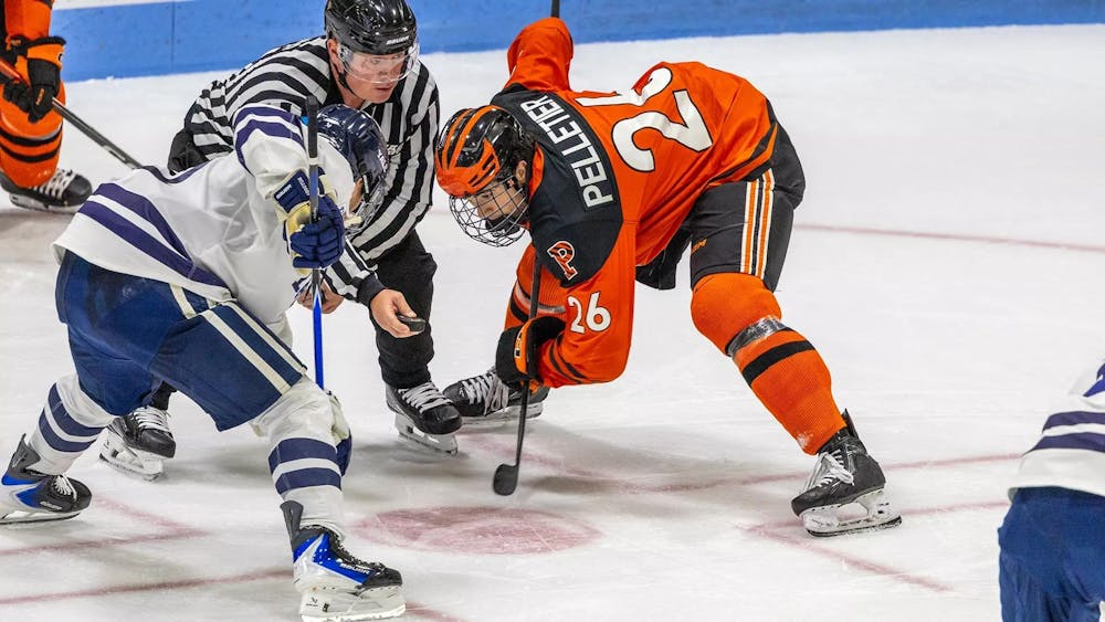 Two hockey players, one in a white and blue jersey and one in an orange and black jersey, do a face-off on the rink.