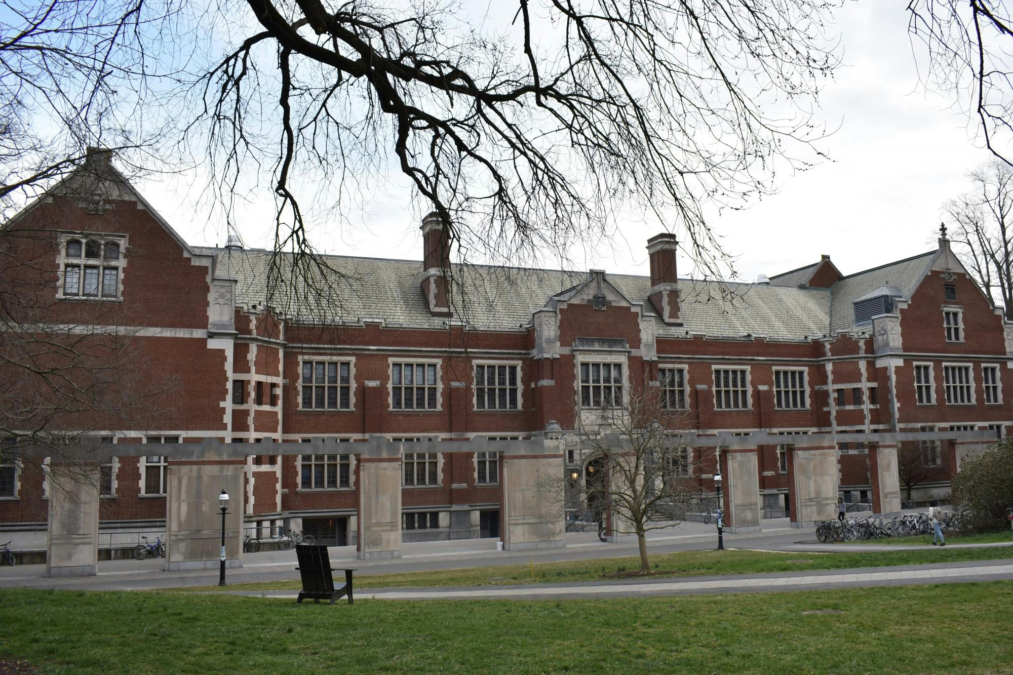 A far view of the front face of the Frist Campus Center from the North Lawn. Pillars partially obstruct the building, which is made of red brick with a gray tiled roof and concrete window frames.