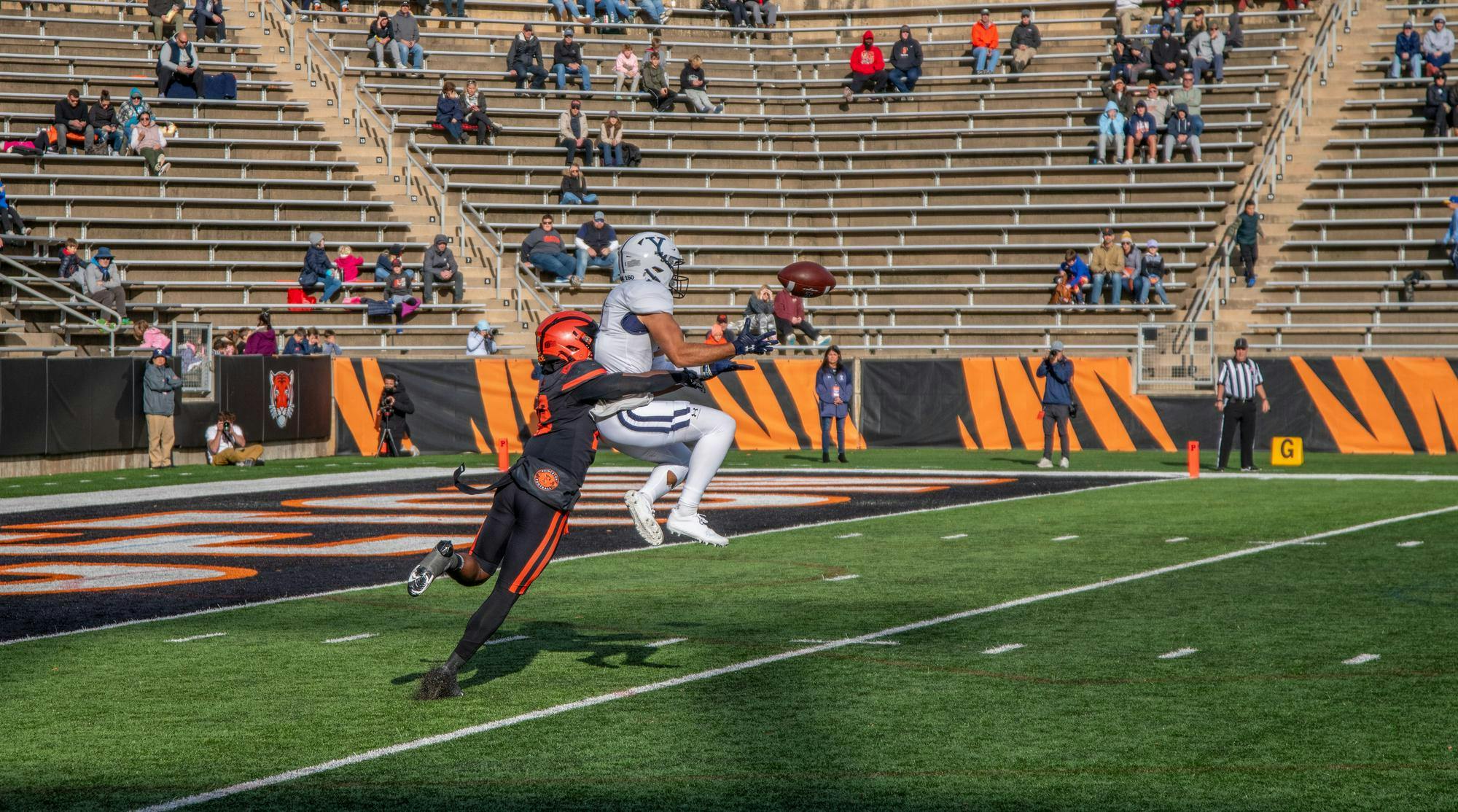 A football player in Yale attire attempts to intercept the football while getting tackled by a football player in Princeton attire.