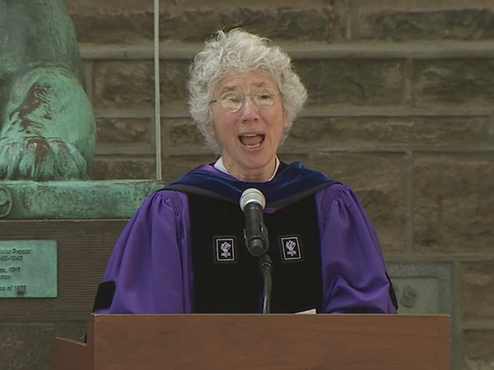 A woman with grey hair wears a purple robe while standing in front of a microphone.