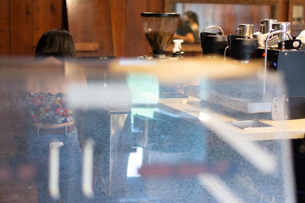 A girl wearing a floral top and jeans sits behind a brown and white coffee shop register. 