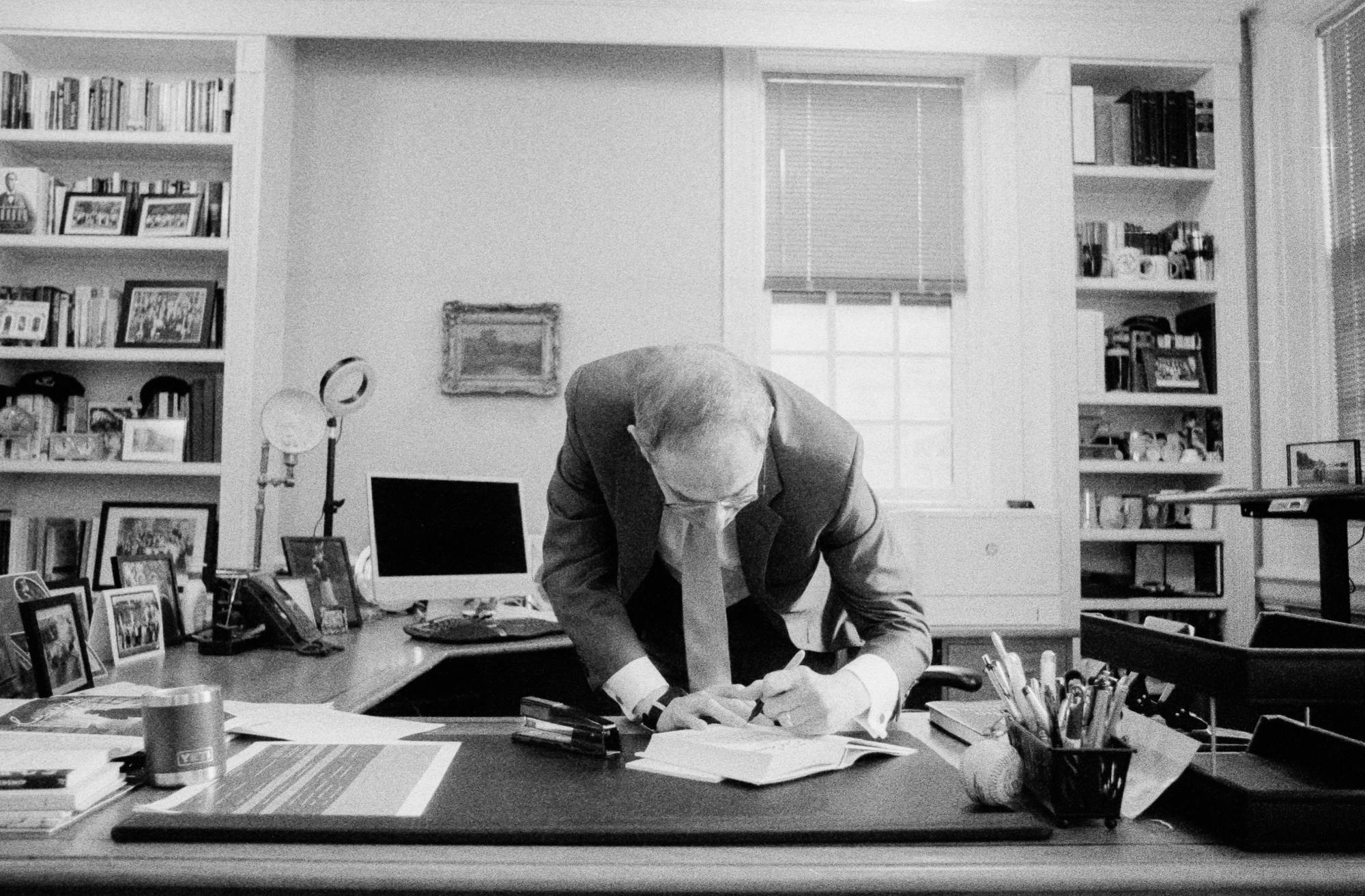 A black and white grainy photo with University President Christopher Eisgruber '83, at the center, bent over his cluttered office desk signing a copy of his new book. 