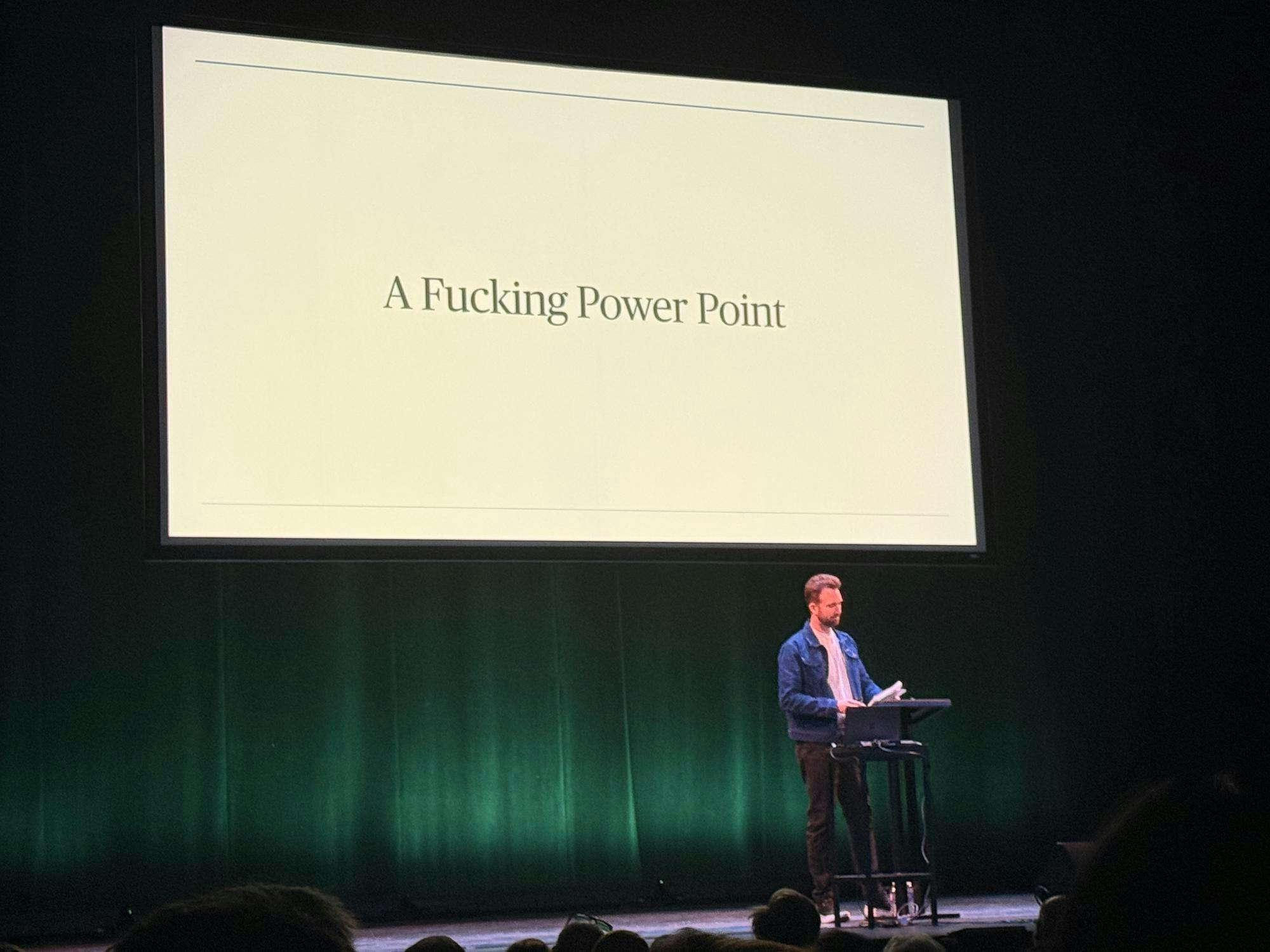 A white man in a blue jacket with a white shirt and brown pants stands at a black podium. There is a white screen that has a presentation presented on it. The presentation reads "A Fucking Power Point" in black font.