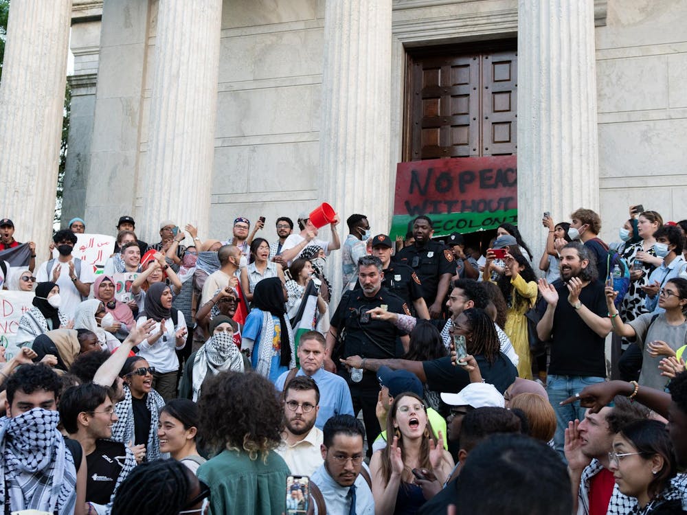 Protestors yelling on the steps of a neo-classical building holding signs in support of the pro-Palestine movement with University Department of Public Safety officers in the middle of the crowd. 