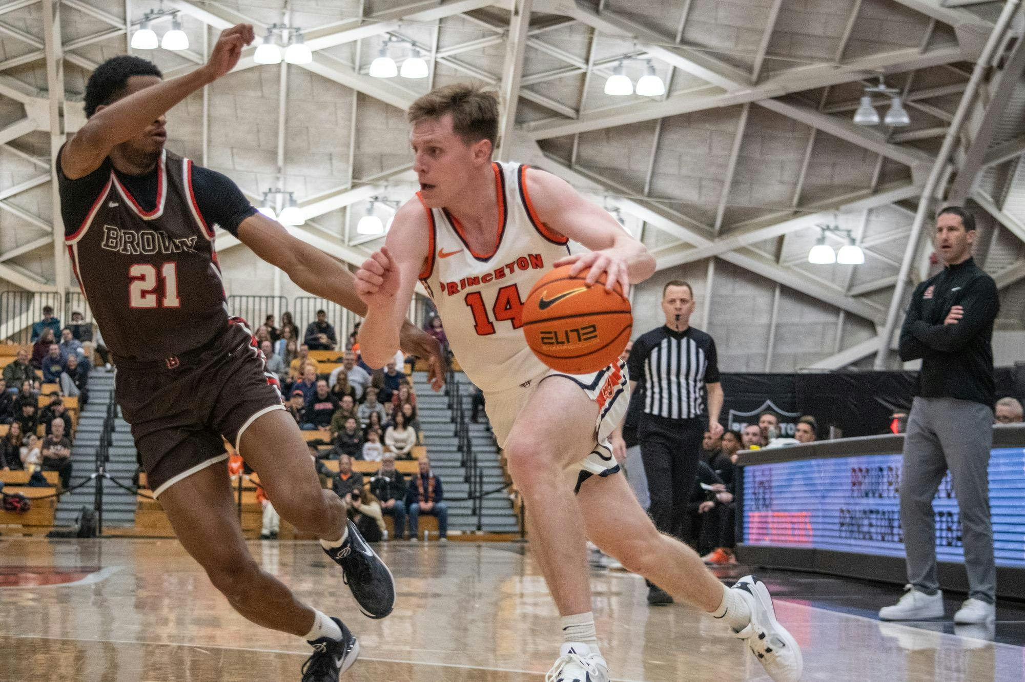 A man shooting a jump shot over a defender at a basketball game while the crowd watches on. 