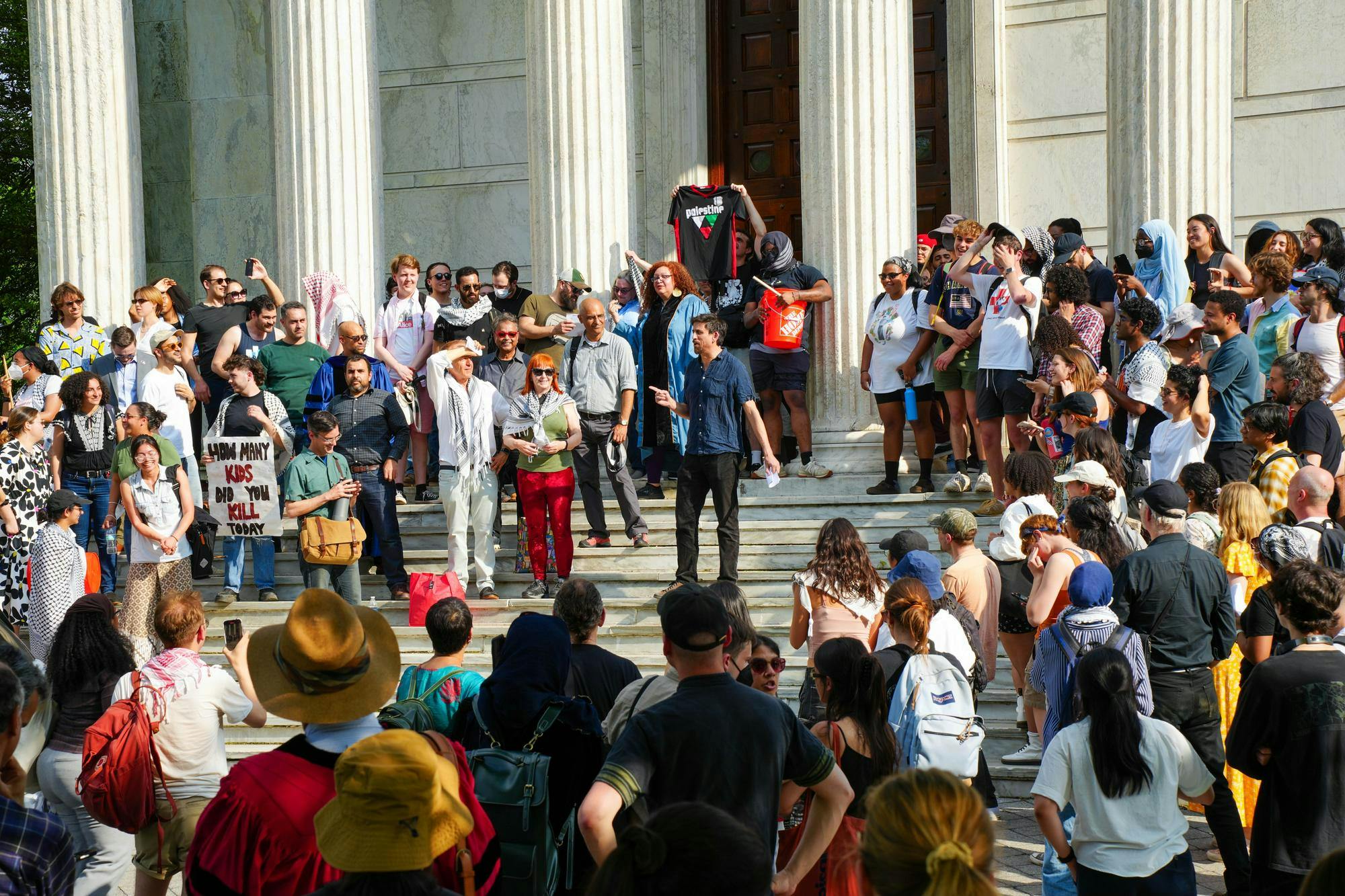 A large group of people congregate outside of a white marble building with pillars. Some carry signs protesting the war in Gaza. 