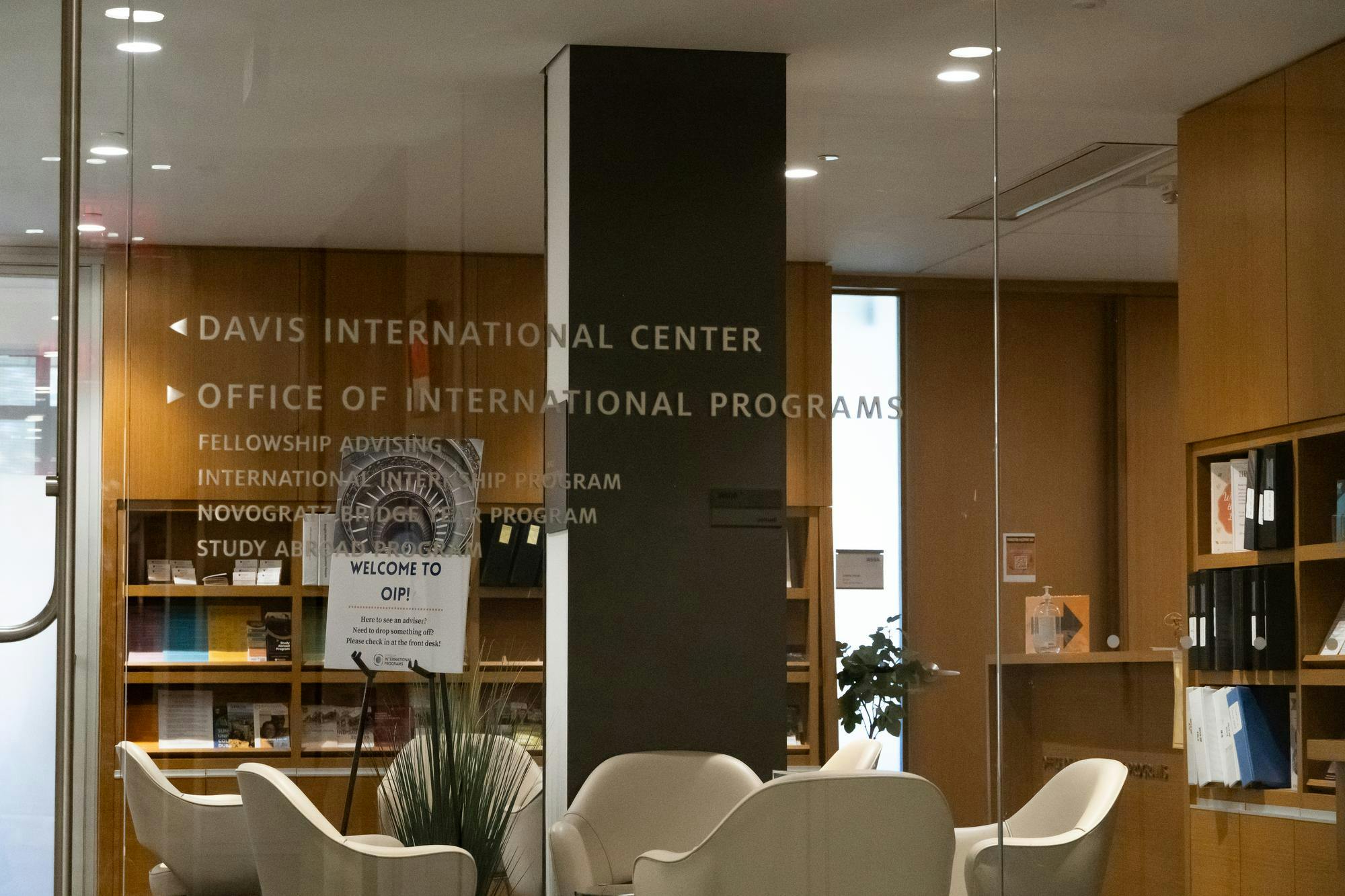 Glass wall reveals an office with wood paneling and white chairs. A black column comes down from the ceiling. There is also a plant. 