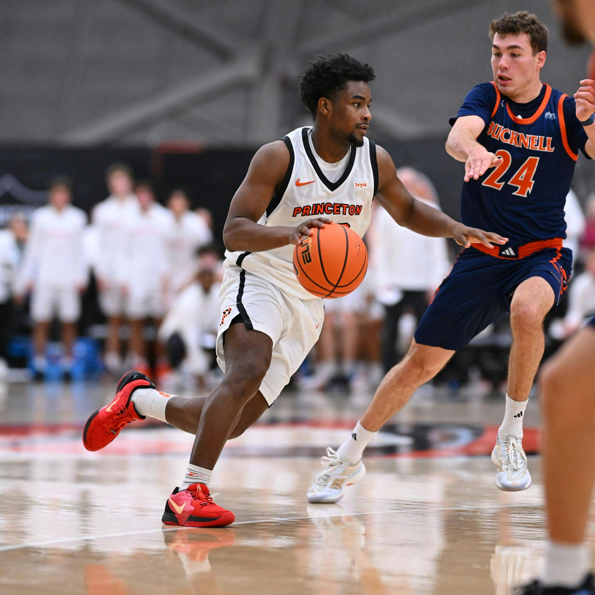 Princeton basketball player in white jersey dribbles basketball past player in navy blue jersey.