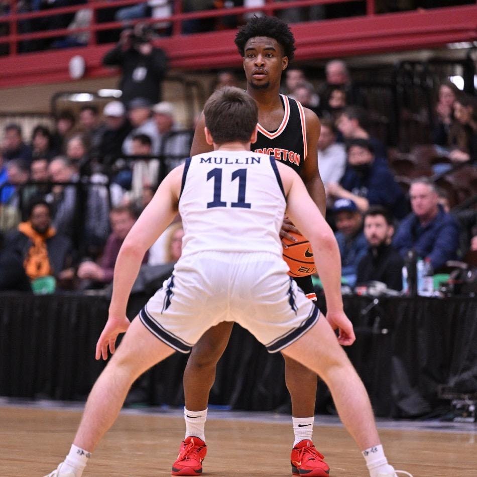 A man stands with a basketball in his hand. He is being guarded by another player. 