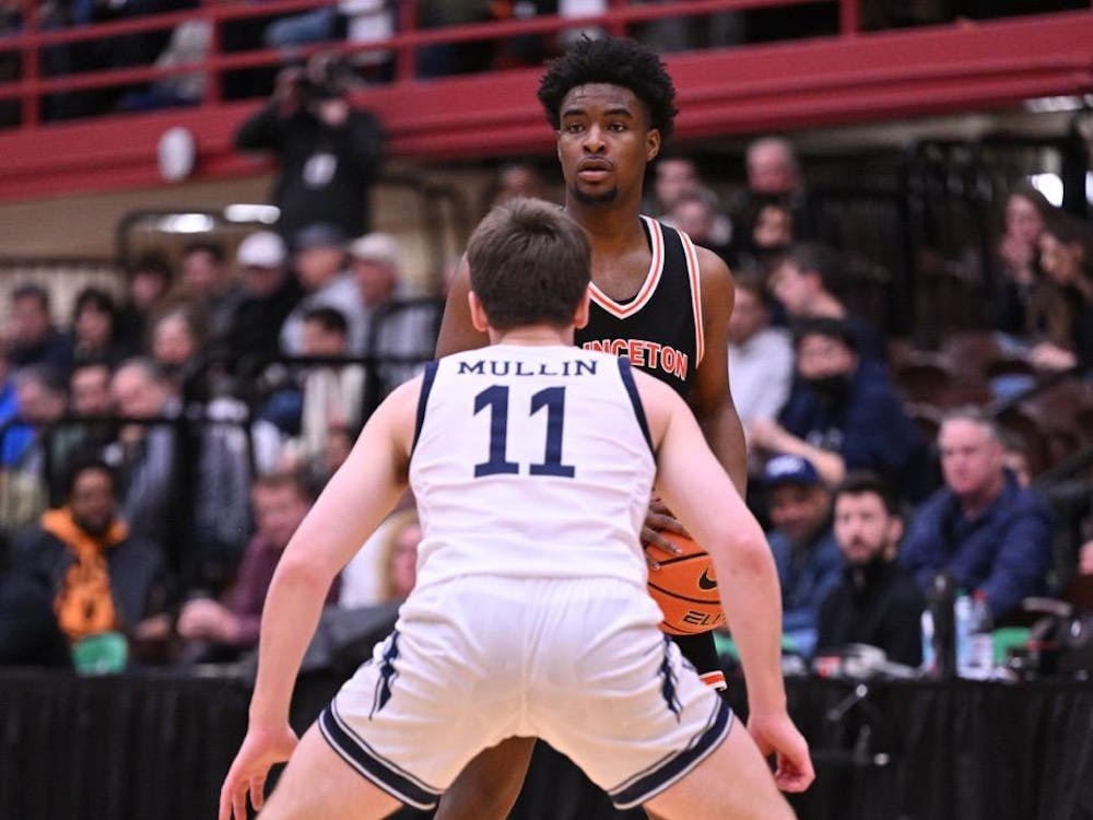 A man stands with a basketball in his hand. He is being guarded by another player.