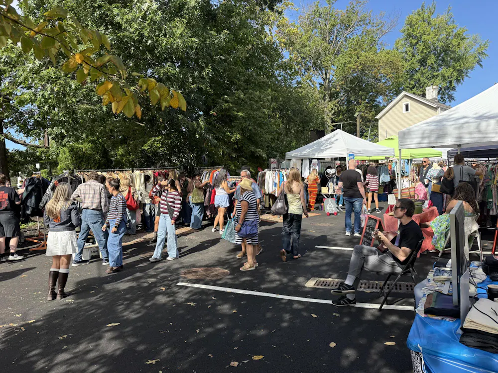 Multiple people are walking around in a parking lot with trees in the background, looking at booths of clothes on a sunny day.