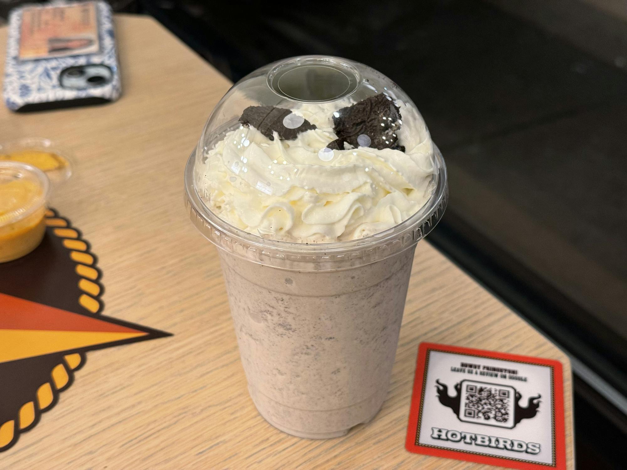 A clear plastic cup has a light brown chunky liquid in it. There is white whipped cream in a spiral pattern on the top of the drink. There are small black oreo cookie pieces on top of the whipped cream. There is a transparent half-dome lid on top of the drink cup.