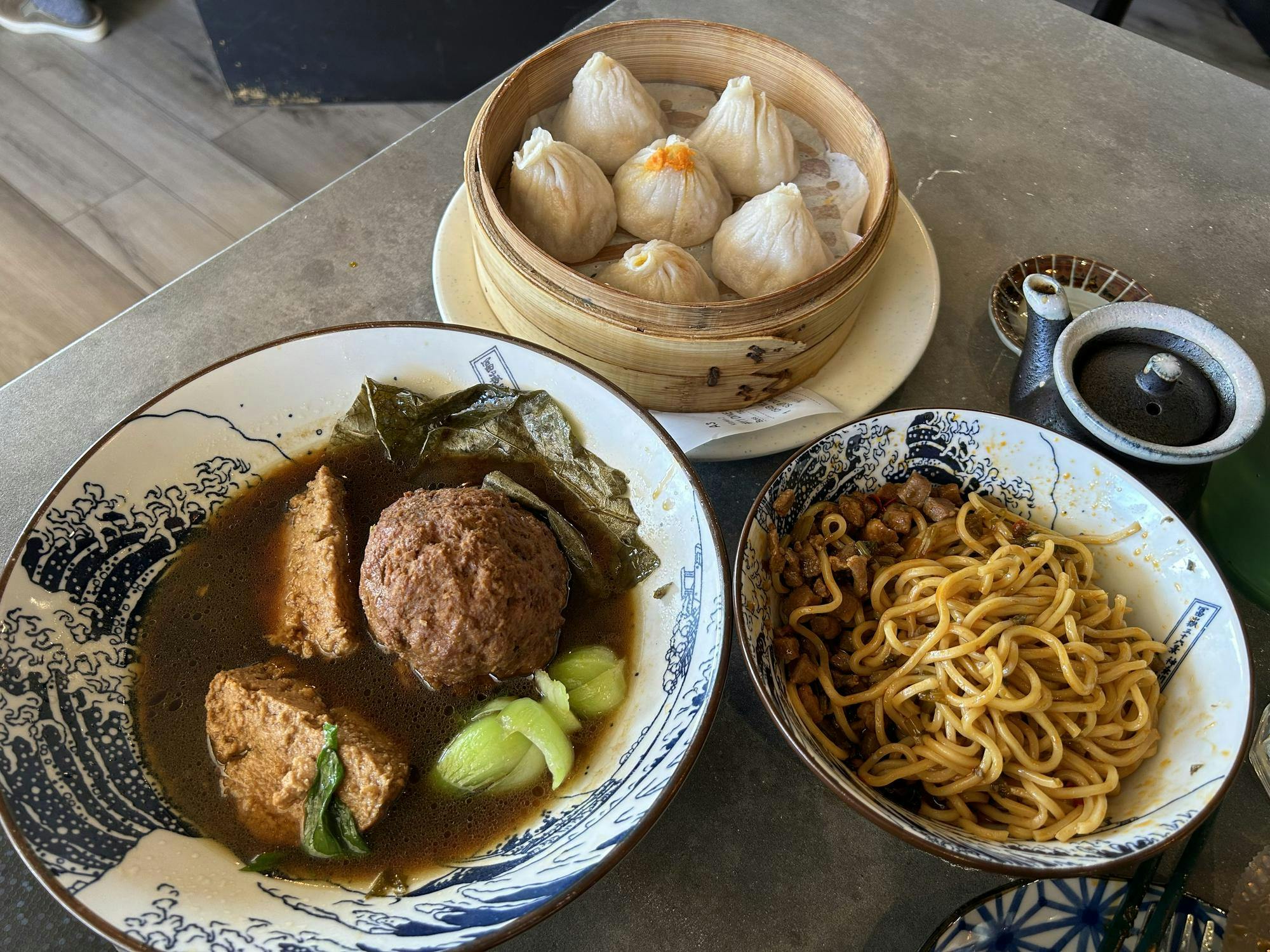 Three dishes, soup dumplings, scallion oil noodles, and jumbo meatballs, sit on a table.