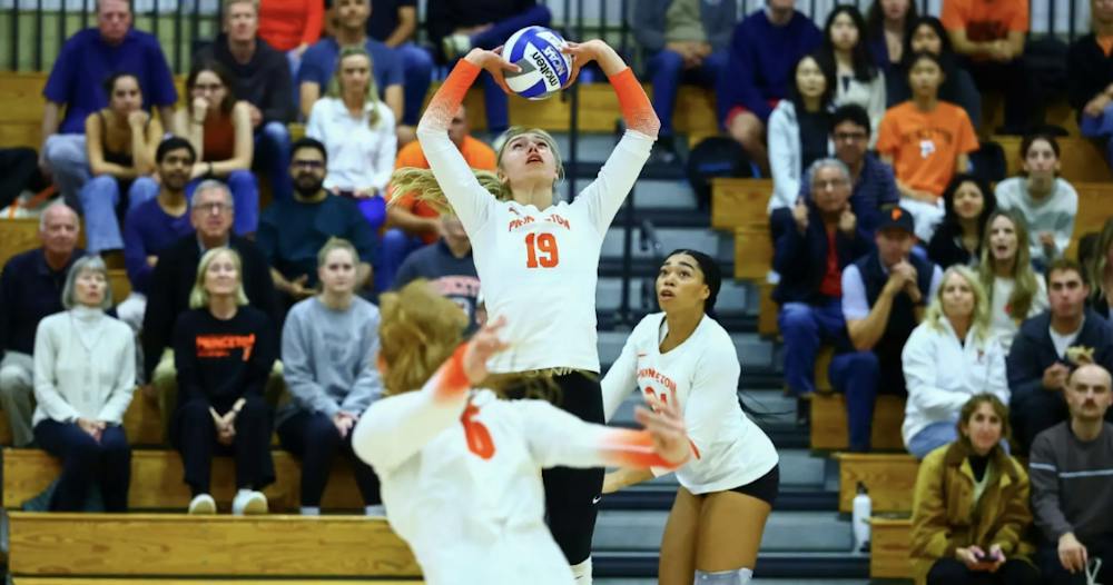 A volleyball player in a white and orange jersey hoisting a volleyball for another player in a white and orange jersey.