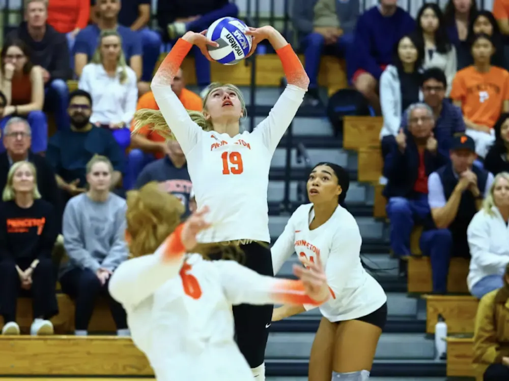 A volleyball player in a white and orange jersey hoisting a volleyball for another player in a white and orange jersey.