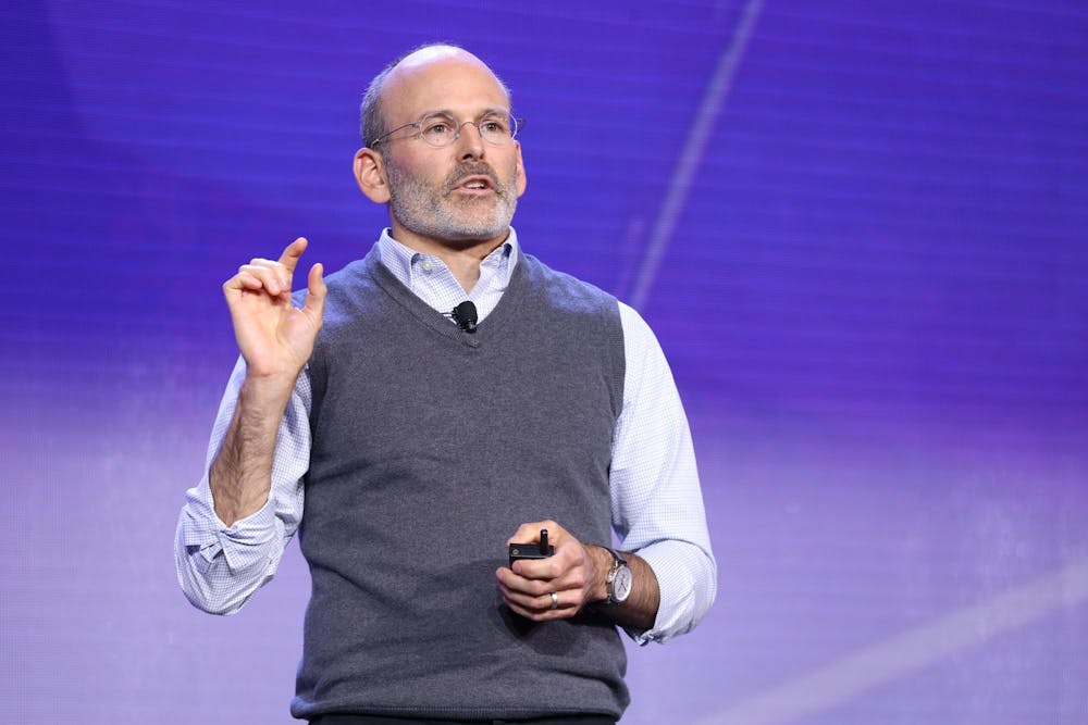 A man wearing a button-up shirt and sweater vest gives a speech on a stage.