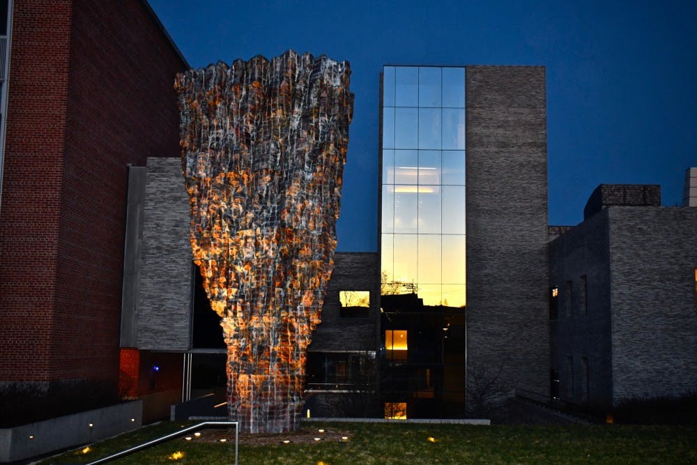 A grey and brick building with reflective window at dusk sits behind a large abstract grey and orange sculpture.