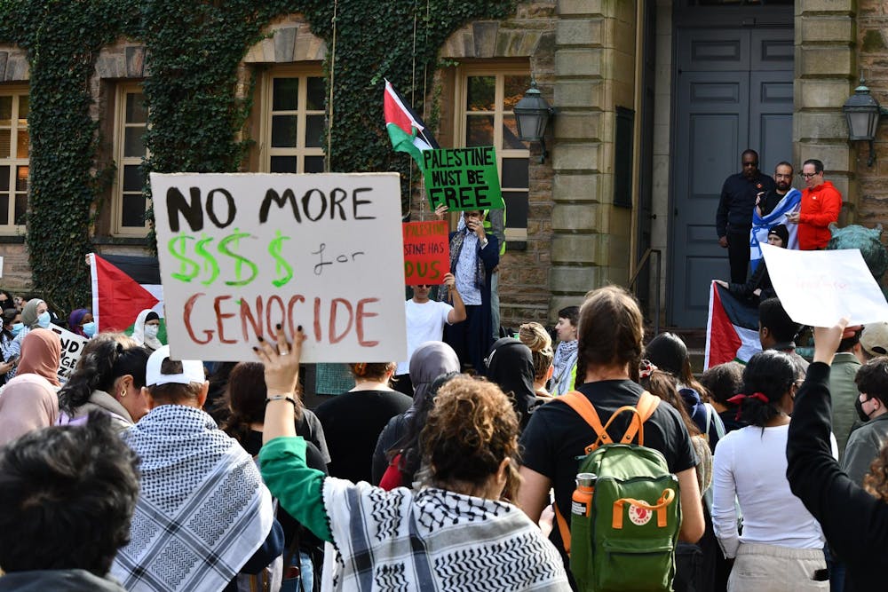 Protestor in front of Nassau Hall, with their back to the camera, wearing a keffiyeh and holding up a sign that reads "No more $$$$ for genocide."