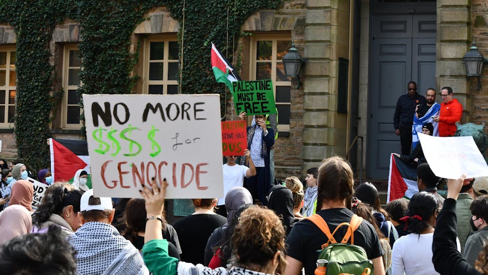 Protestor in front of Nassau Hall, with their back to the camera, wearing a keffiyeh and holding up a sign that reads "No more $$$$ for genocide."