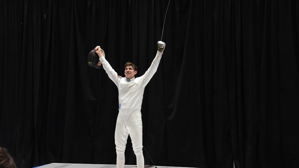 Man in white fencing suit raises arms and épée.