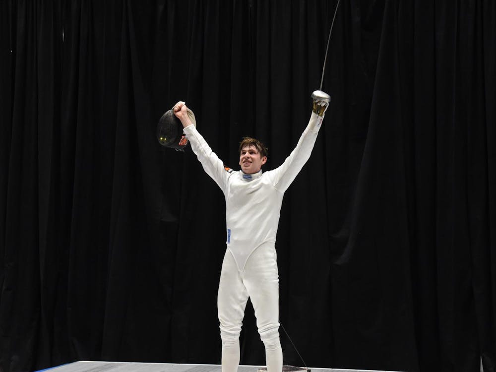 Man in white fencing suit raises arms and épée.