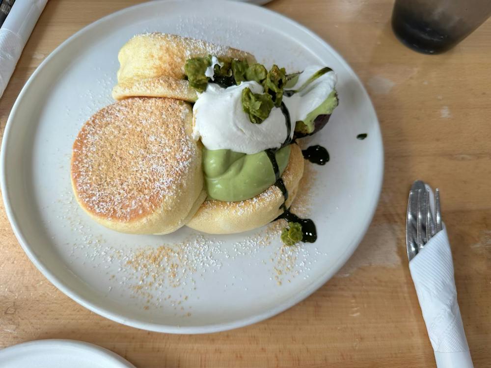 There is a white plate on a light brown table. On the plate, there are three light brown pancakes. The pancakes are covered in white cream, green cream, dark brown sauce, and powdered sugar.