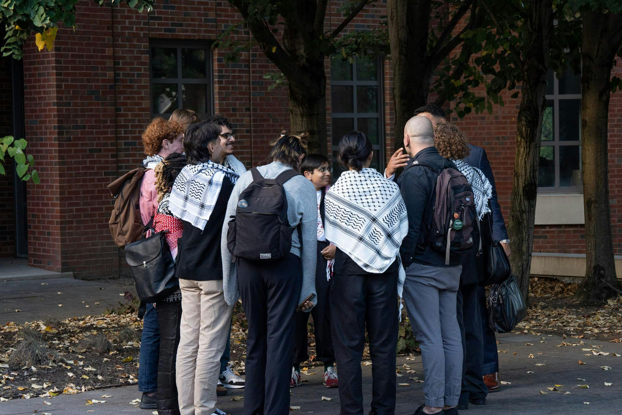 A small group of people wearing keffiyehs stand in a circle outside of a large brick building.