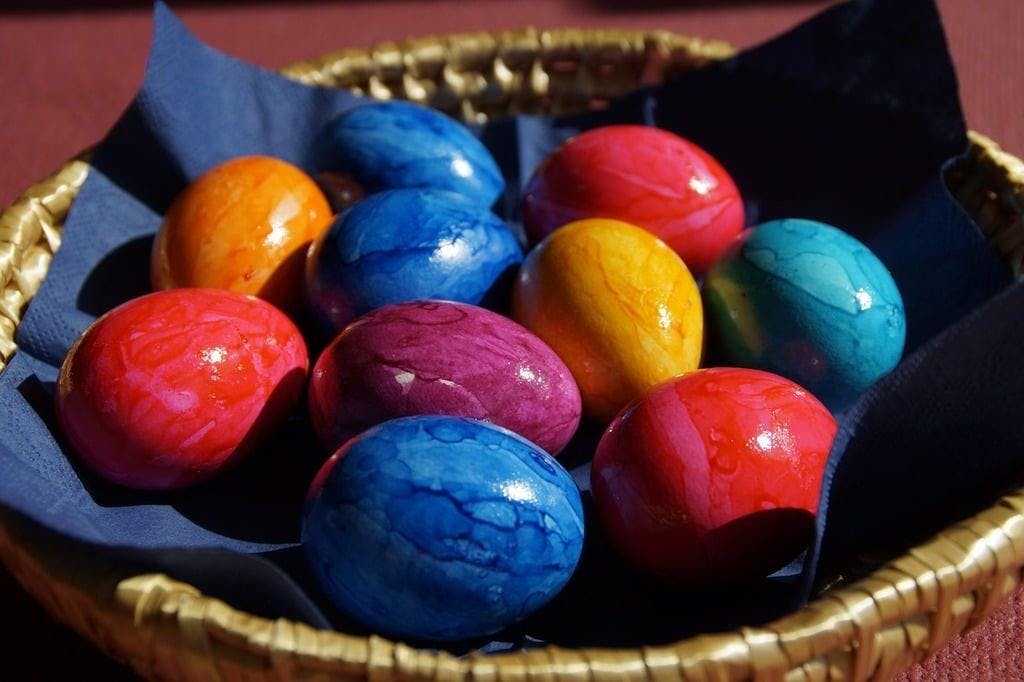 Blue, orange, red, and purple painted eggs on a blue napkin in a brown basket with a reddish-brown background.