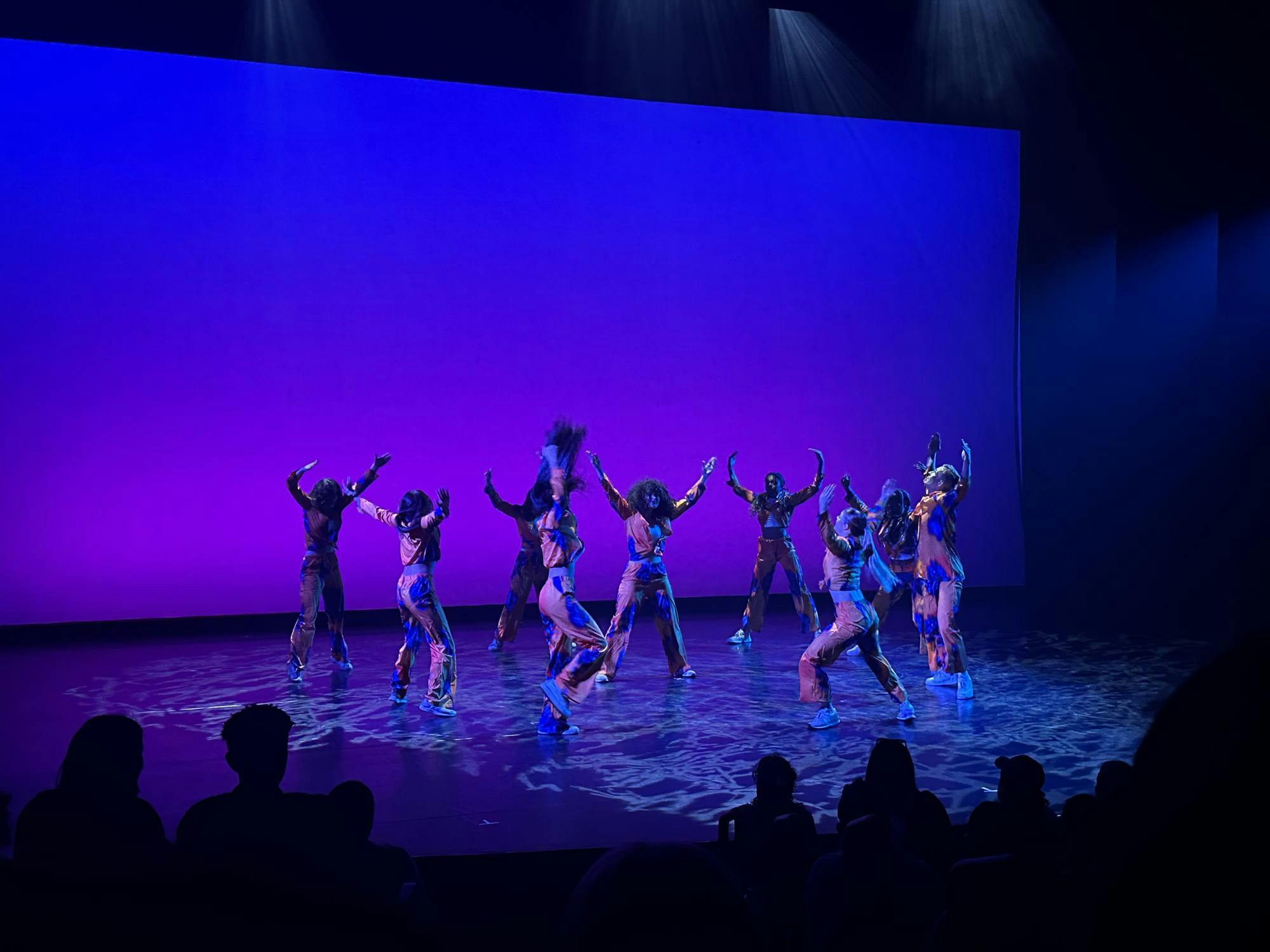 Dancers mid-movement gather in a circle on a black stage illuminated by purple lighting.