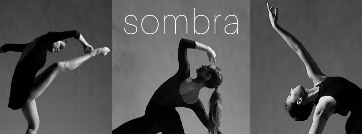 A female dancer with a long ponytail stands in a pose with her back arch and arm lifted over her head, gazing up toward the sky. The word "sombra" is written in white above her head, and two other dancers are partially visible to either side of her. The photograph is in black and white. 