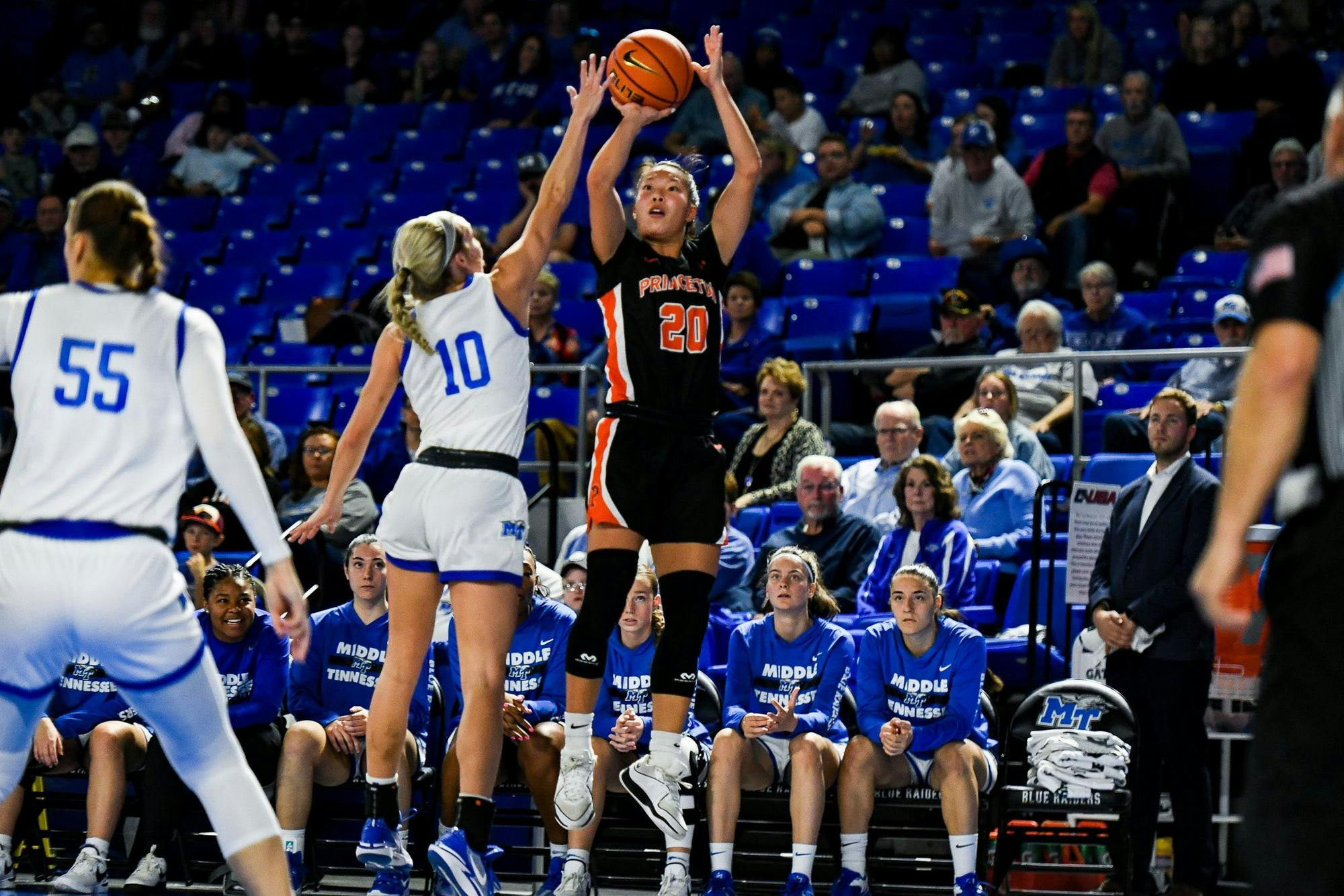 A woman wearing a black jersey and black shorts shoots a basketball over a defender wearing a white jersey and shorts as a crowd watches on. 