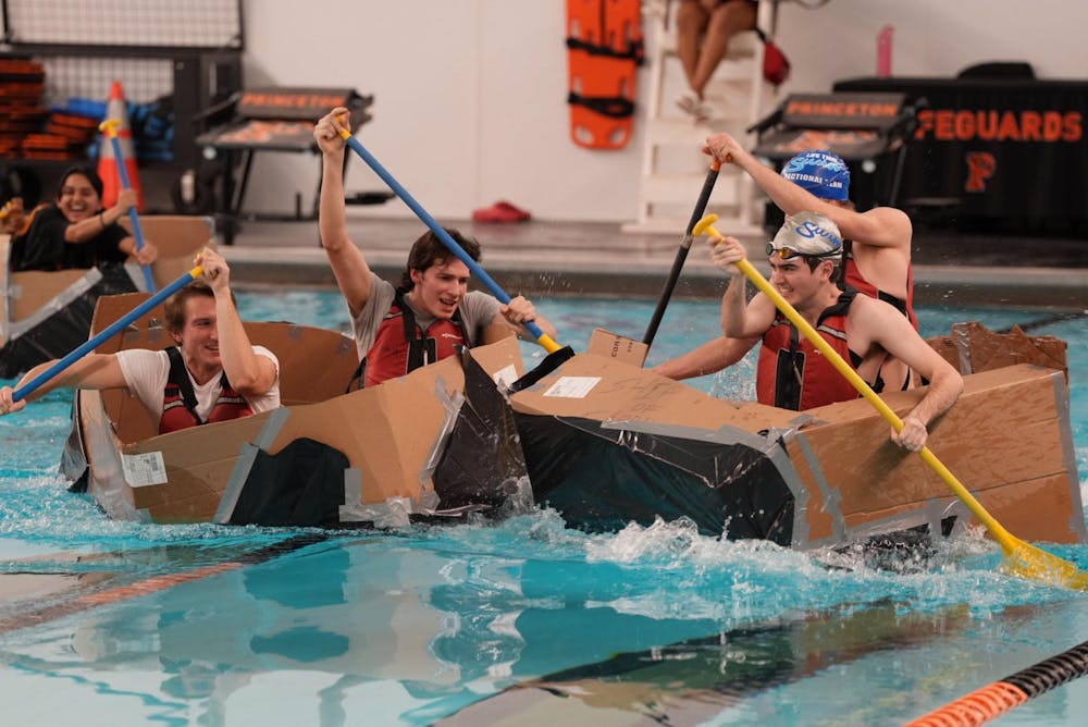 Four men paddling across a swimming pool in homemade cardboard boats.
