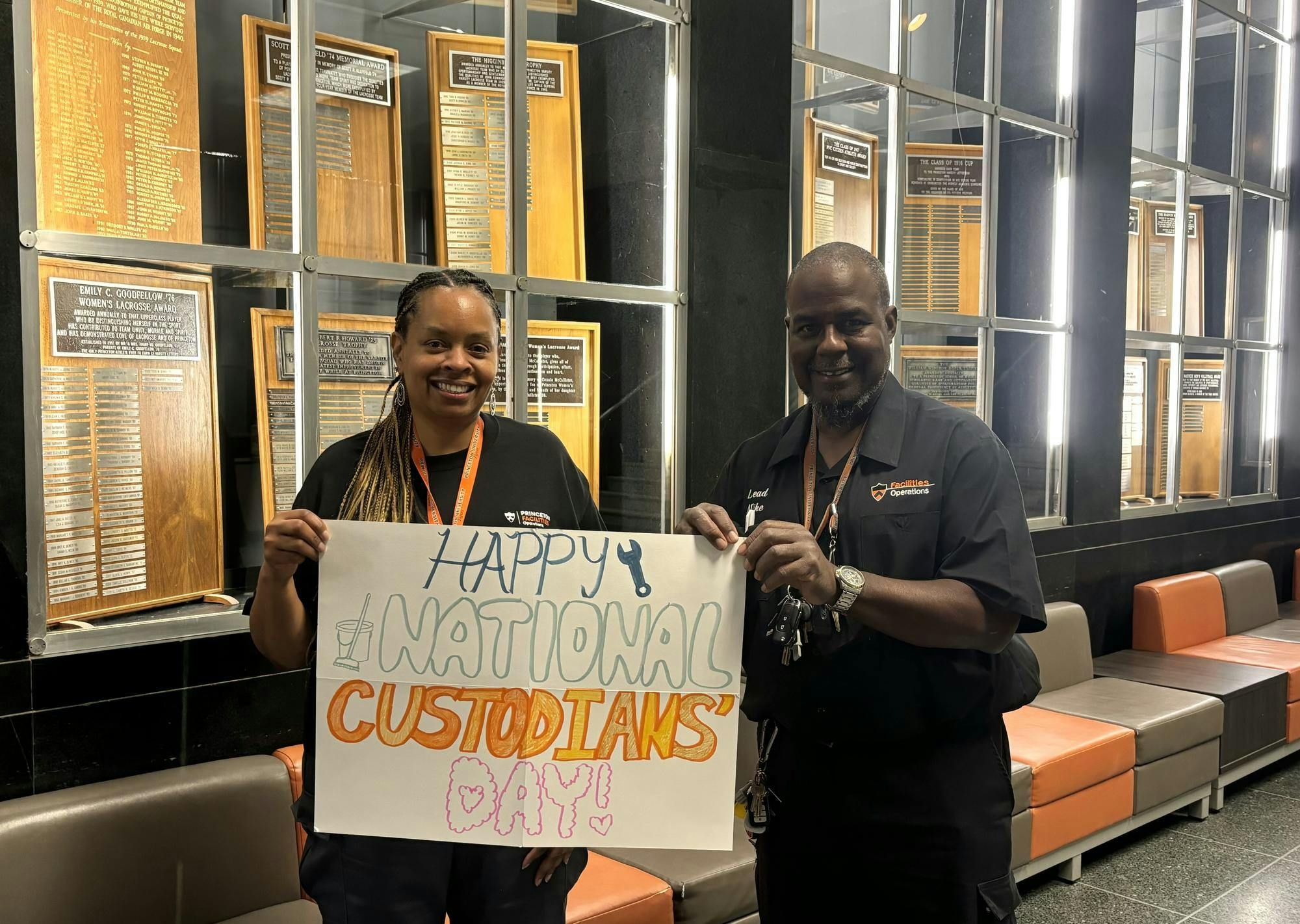 Two smiling people wearing black uniforms with orange and white text hold a large white sign reading "Happy National Custodians Day." 