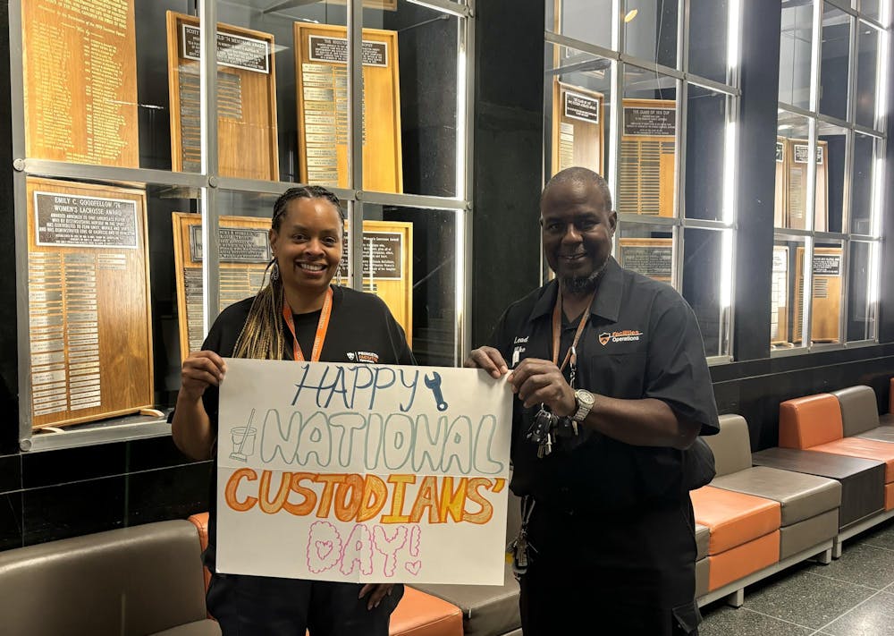 Two smiling people wearing black uniforms with orange and white text hold a large white sign reading "Happy National Custodians Day." 