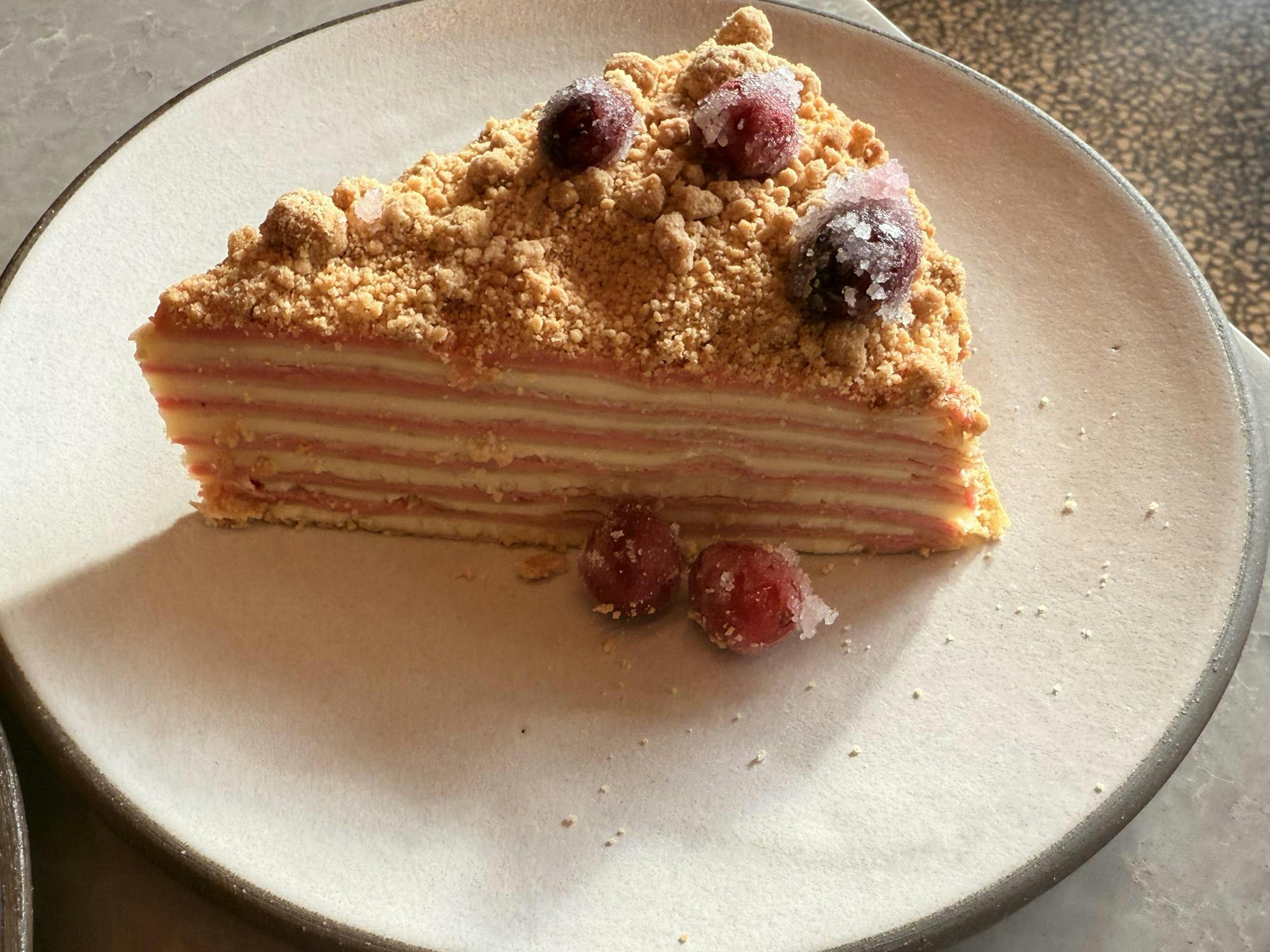 A multi-layered crepe cake topped with cake crumble and sugar-coated cranberries served on a white plate.