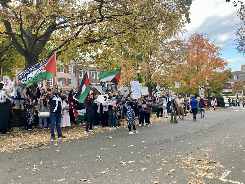 A crowd of people stands on the sidewalk and in the street, holding up posters and Palestinian flags.