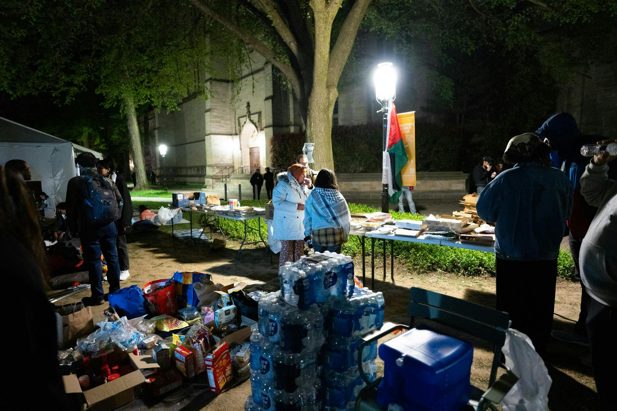Piles of water bottles and food, two people stand under a light fixture in the center of the frame