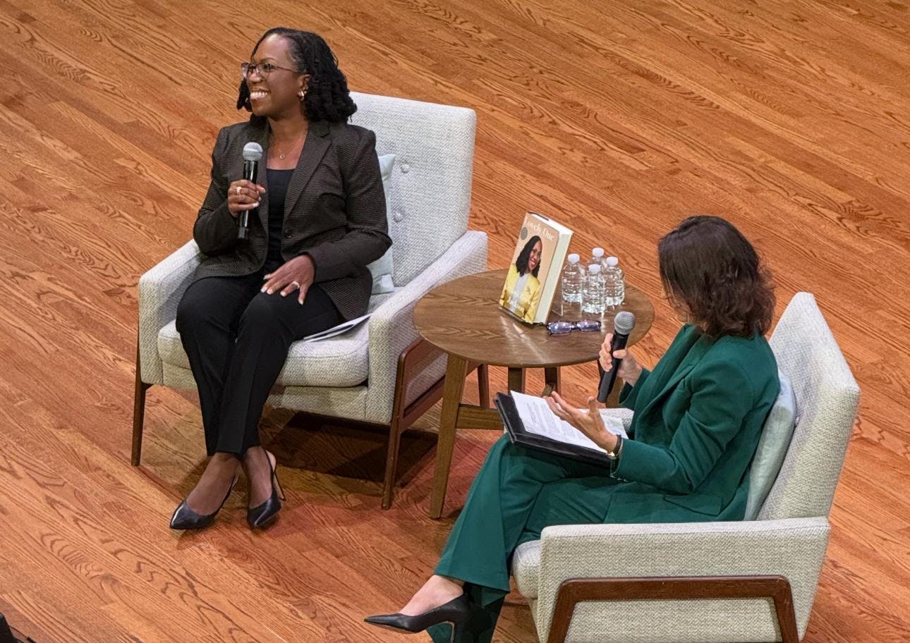 Two women are seated in chairs, holding microphones, speaking to a large audience inside an auditorium. 