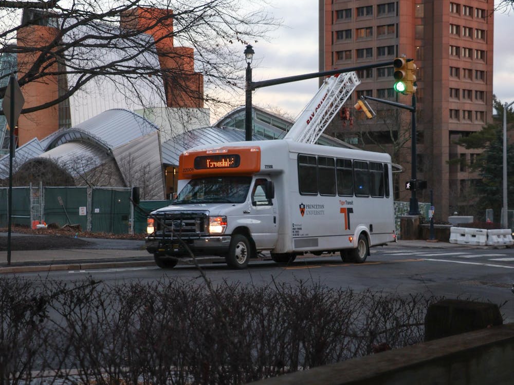 A mostly white bus with orange text and a Princeton University logo drives up the far side of the road, passing a large silver building.