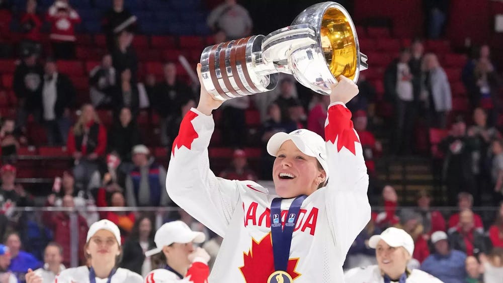 A woman with a medal around her neck holds up a trophy in the air on an ice hockey rink. 