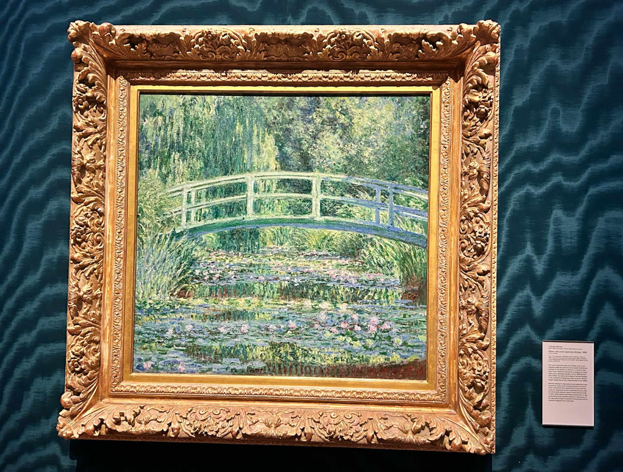 A painting of a bridge crossing over clusters of water lilies in an ornate brown frame. 