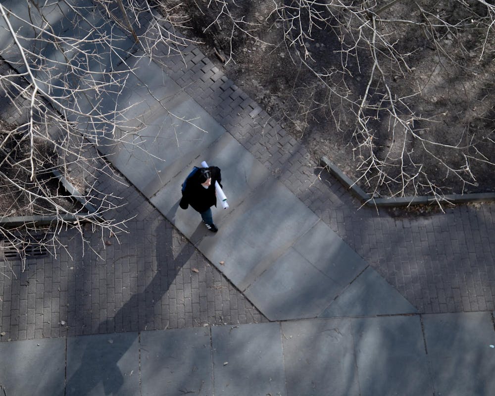Overhead view of student walking on sidewalk holding posters; tree branches visible. 