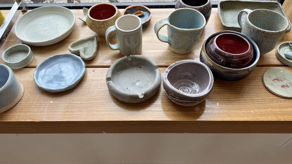 An assortment of ceramic mugs, bowls, and containers on a wooden shelf by a window.