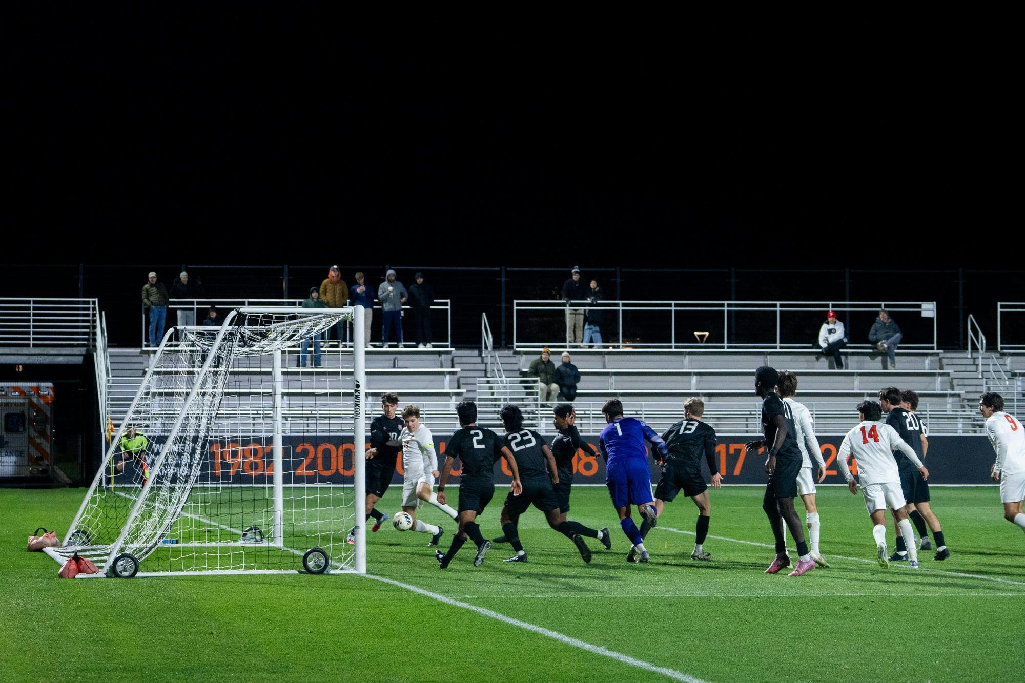 Soccer players crowd in front of the goal at night as a white uniformed player takes a close shot, with defenders and the goalkeeper trying to stop it.