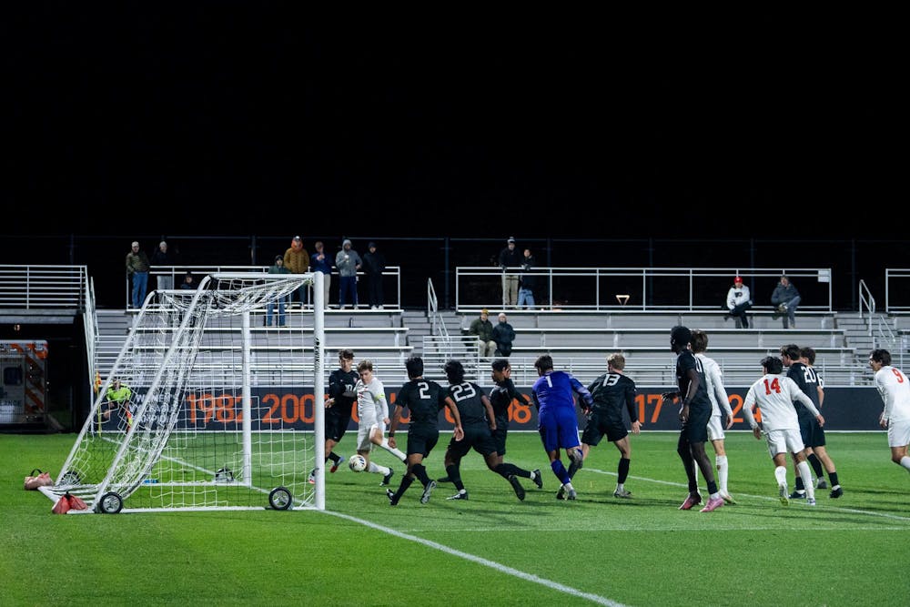 Soccer players crowd in front of the goal at night as a white uniformed player takes a close shot, with defenders and the goalkeeper trying to stop it.