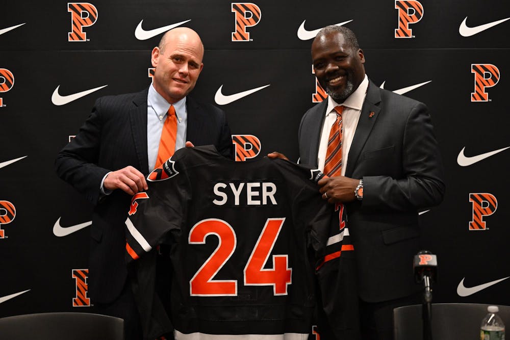 Two men posing with a hockey jersey at a press conference with the Nike and Princeton logos displayed in the background 